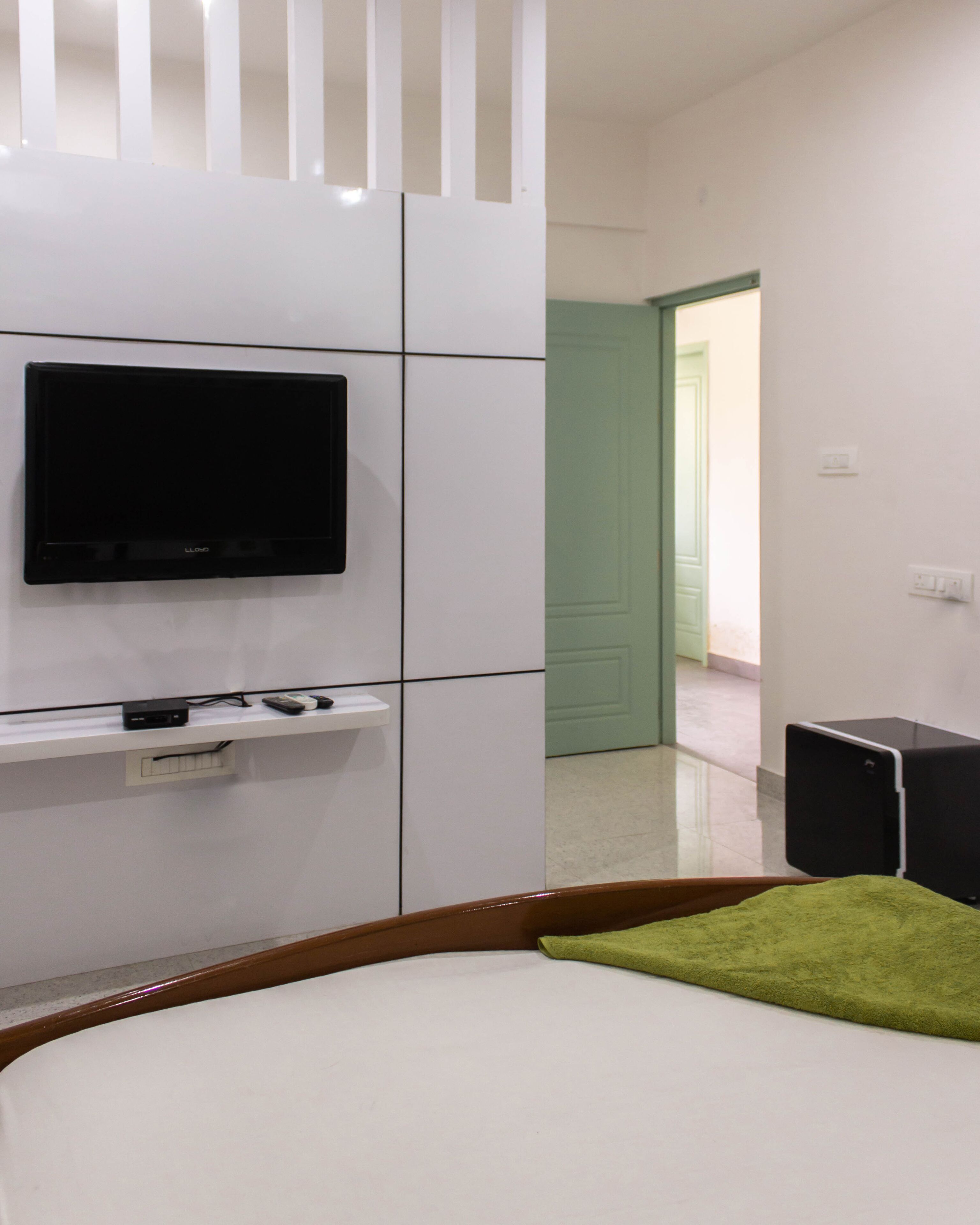A hotel room with a bed, a TV mounted on the wall, a small shelf with a remote control, and a green towel on the bed.
