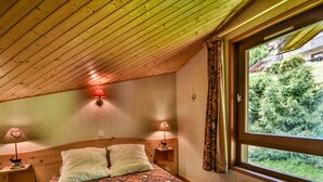 Ein Schlafzimmer mit Holzboden, einem Bett mit gemusterten Textilien, zwei Nachttischlampen und einem Fenster mit Blick auf Grünflächen.