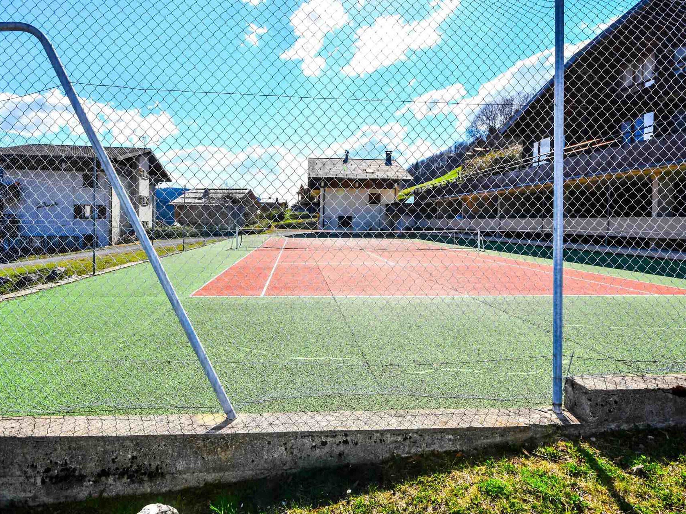 A tennis court enclosed by a chain-link fence with a clear sky and buildings in the background.