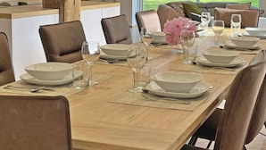 A dining area with a long table set for a meal, surrounded by chairs, with a view of greenery outside.