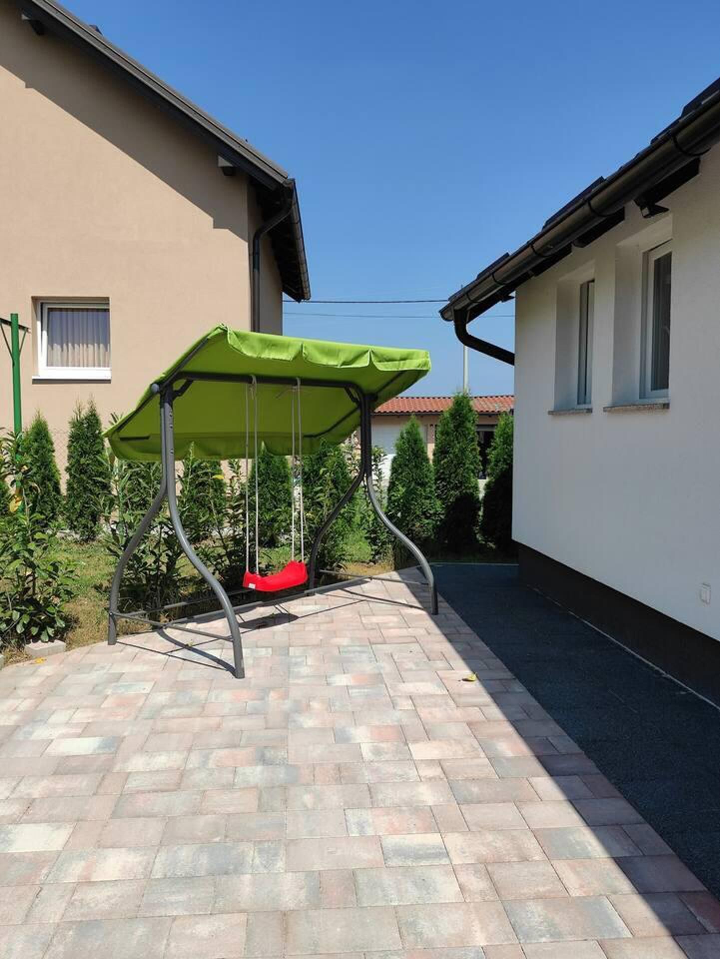 A patio area with a swing set under a green canopy, surrounded by a paved walkway and adjacent buildings.