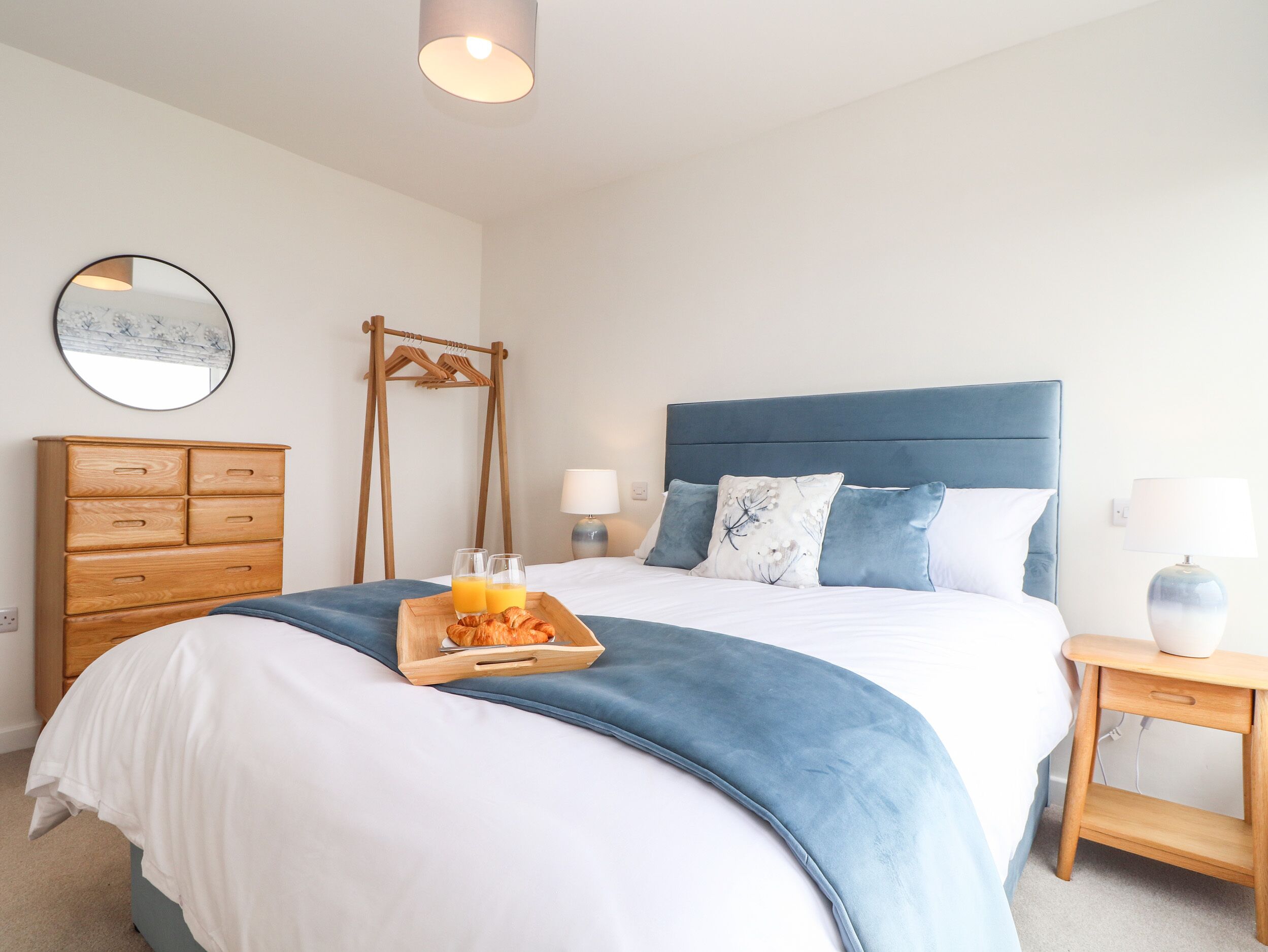 A neatly made bed with a tray of croissants and orange juice, a wooden dresser, a mirror, and a bedside table with a lamp.