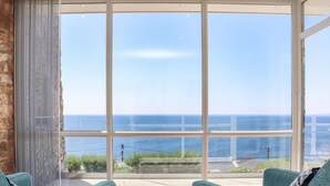 Una habitación con un ventanal grande que ofrece vistas al mar, dos sofás color turquesa y un reposapiés marrón.