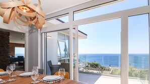 Comedor con mesa de madera dispuesta para cuatro, un ventanal con vistas al océano y una lámpara moderna.
