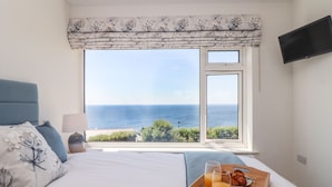 Un dormitorio con un ventanal grande con vista al mar, una cama con almohada floral, una bandeja con alimentos para el desayuno y un televisor en la pared.