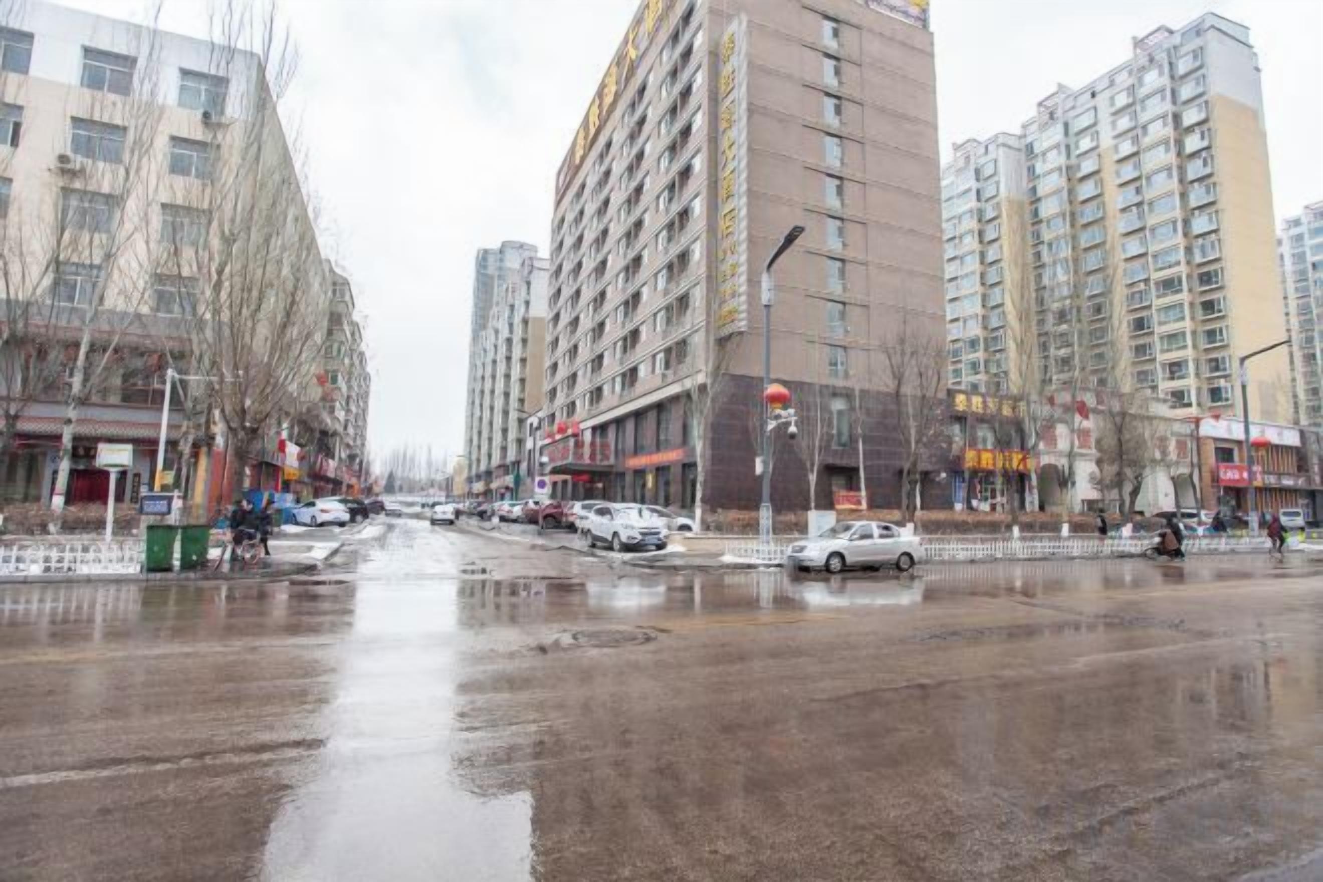 A wet urban street with tall buildings, parked cars, and a clear sky.
