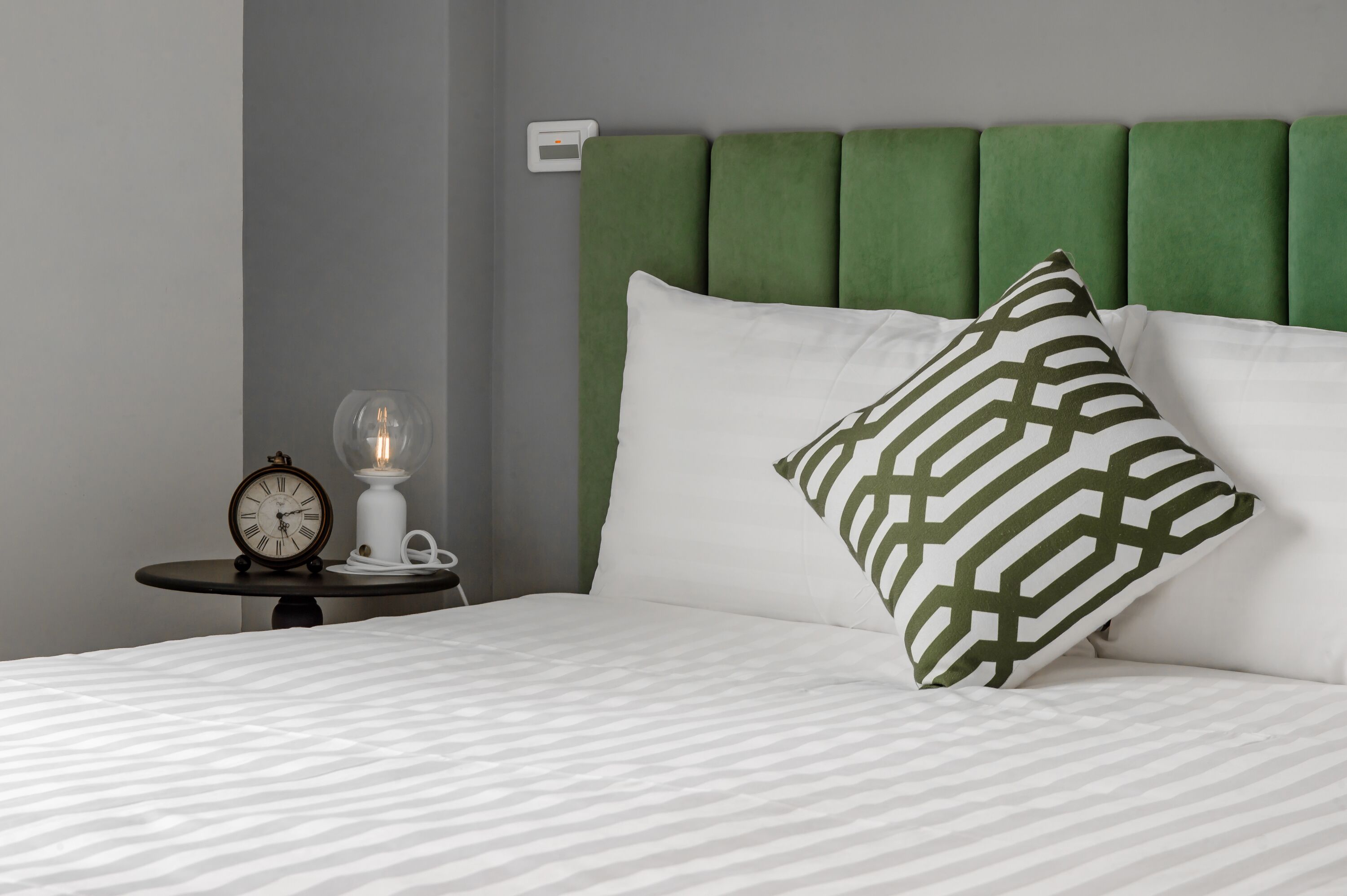 A neatly made bed with a patterned pillow, a bedside table with a clock and a lamp, and a green upholstered headboard.