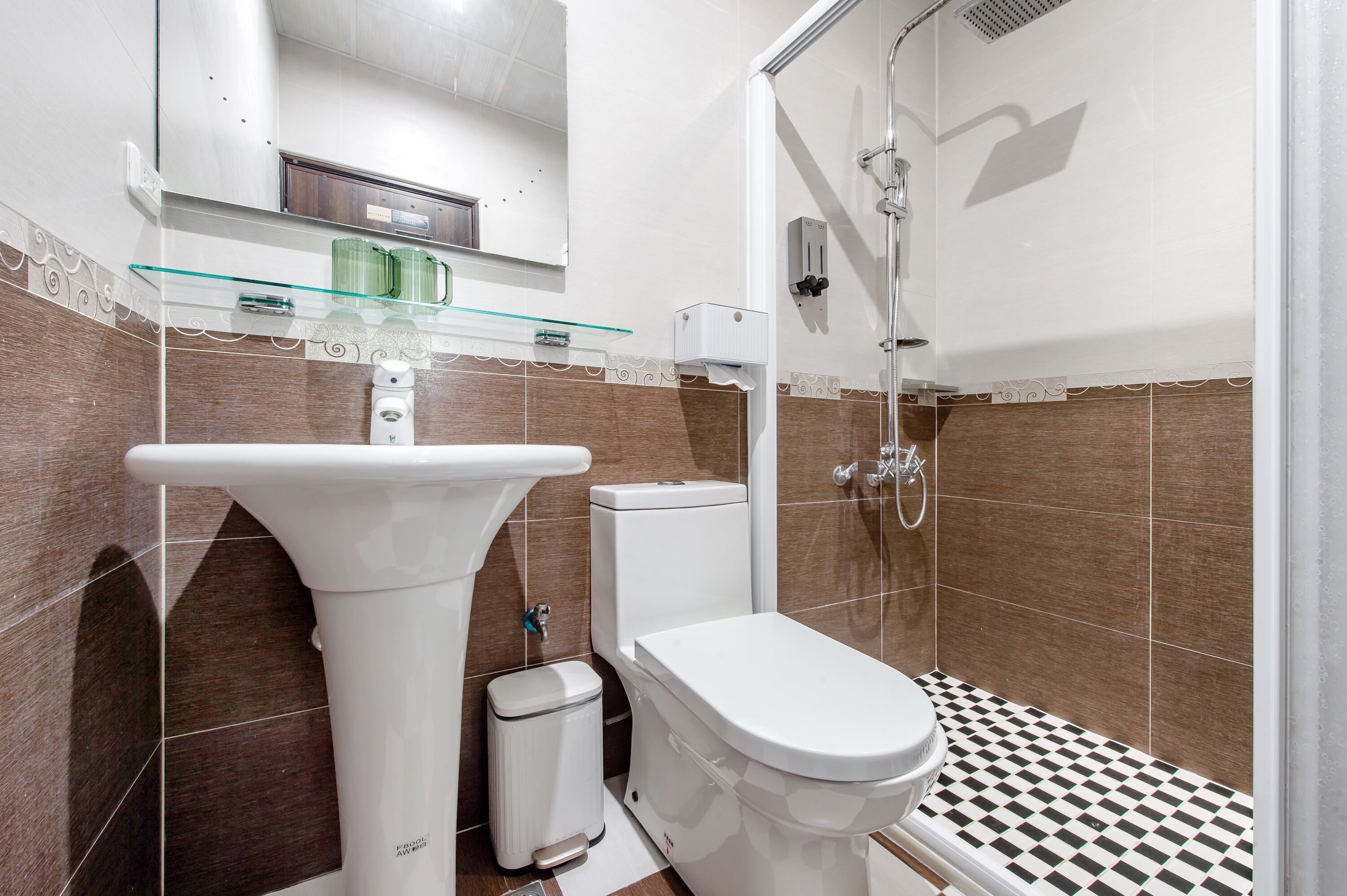 A bathroom with a white sink, toilet, and shower. There is a glass shelf above the sink and a trash bin next to the toilet.
