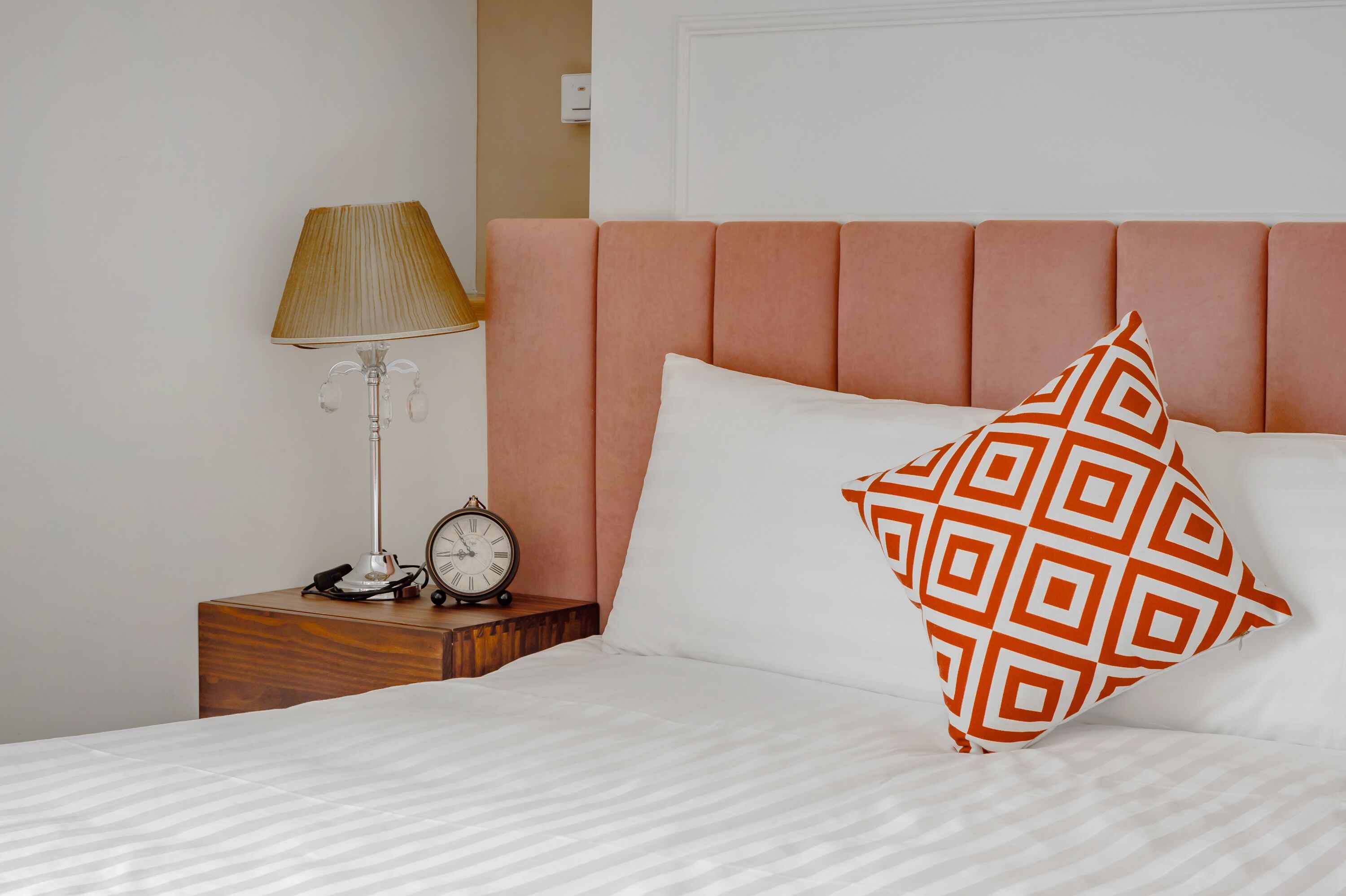 A neatly made bed with a patterned pillow, a bedside lamp, and a clock on a wooden nightstand.