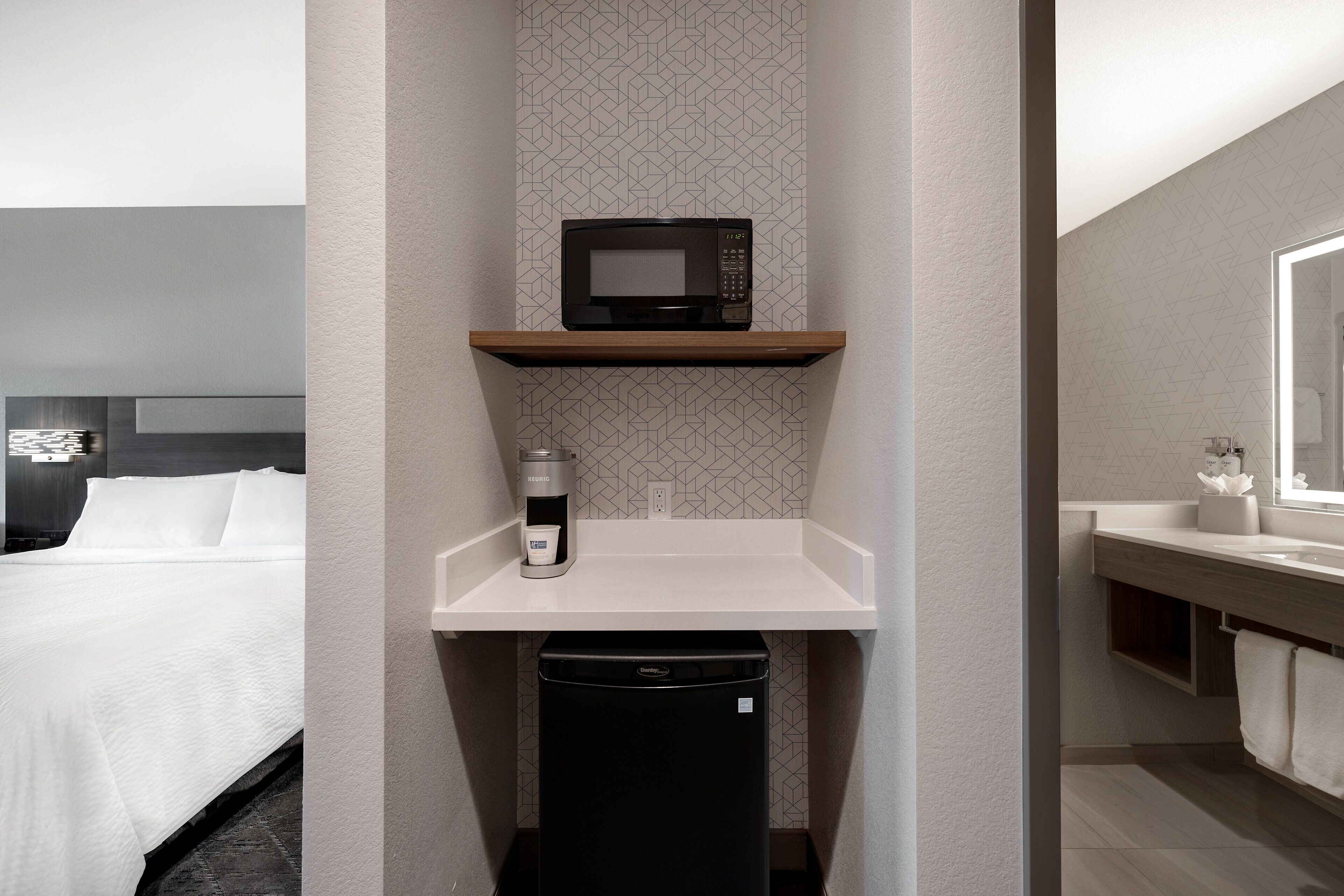 A compact hotel room with a bed, a microwave on a shelf, a coffee maker, and a compact dishwasher.