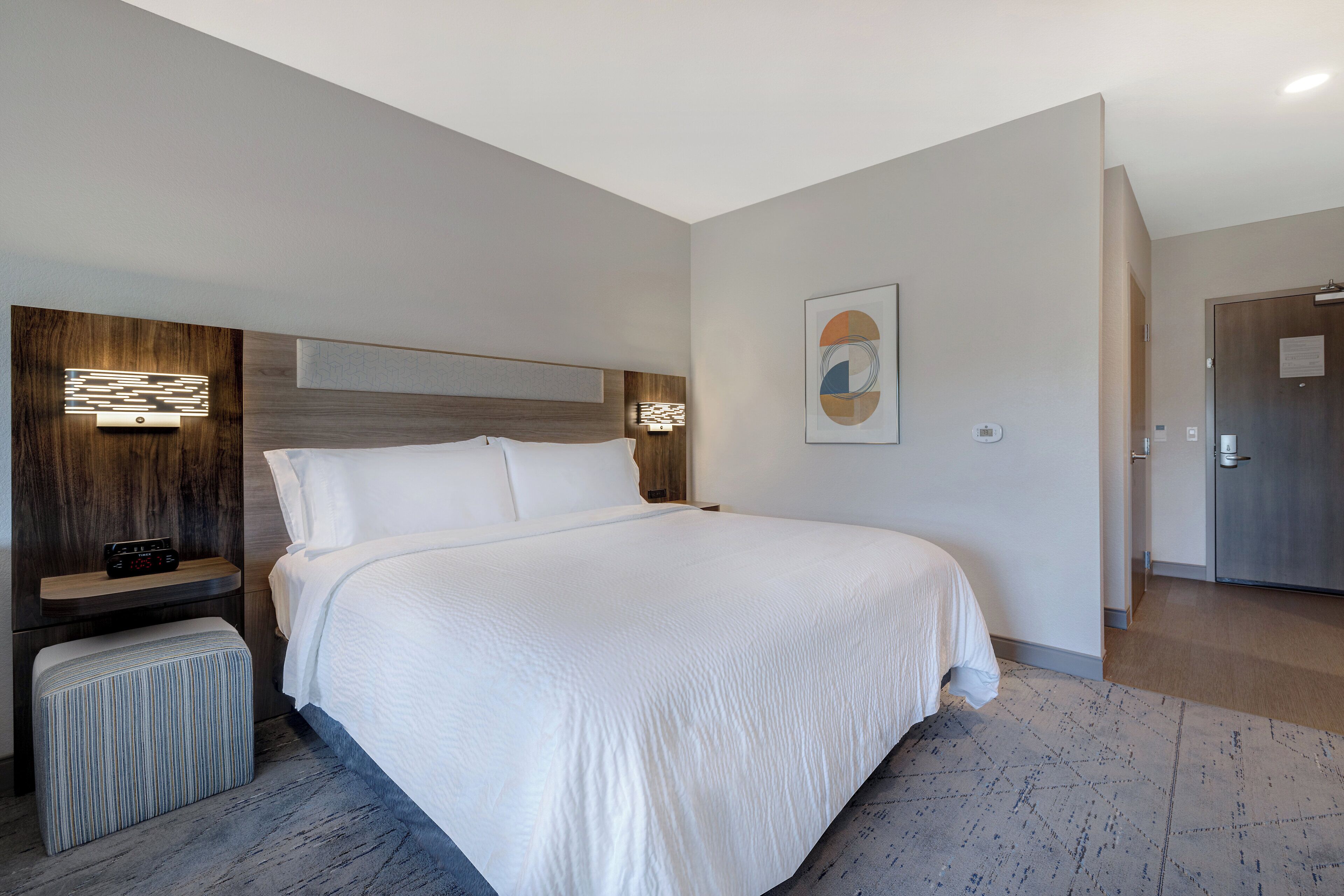 Una habitación de hotel moderna con una cama grande, una mesita de noche, una lámpara de pared y una puerta.