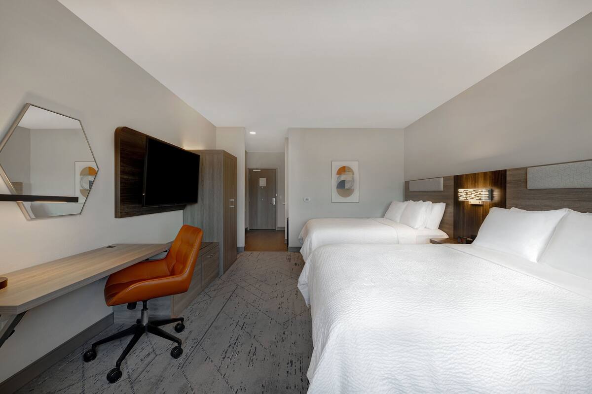 Habitación de hotel con escritorio, una silla de oficina naranja, dos camas, un televisor de pantalla plana y obras de arte en las paredes.