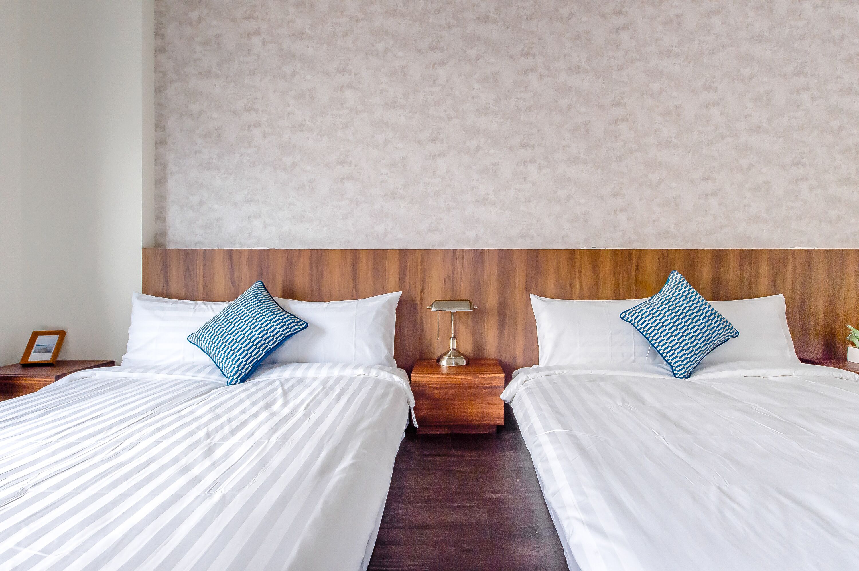 Habitación de hotel con dos camas, cabeceros de madera, ropa de cama blanca y una mesita de noche con lámpara entre las camas.