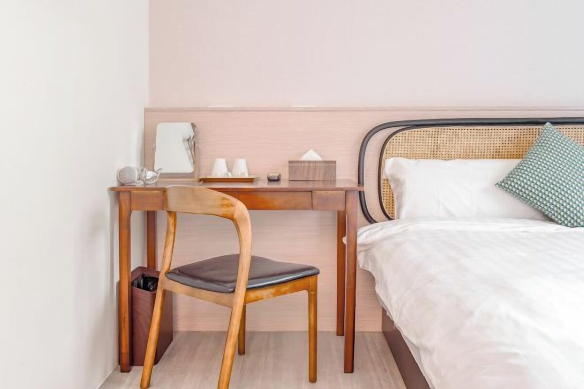 A bedroom with a bed, a wooden desk, a chair, and a bedside table with a lamp and a tissue box.