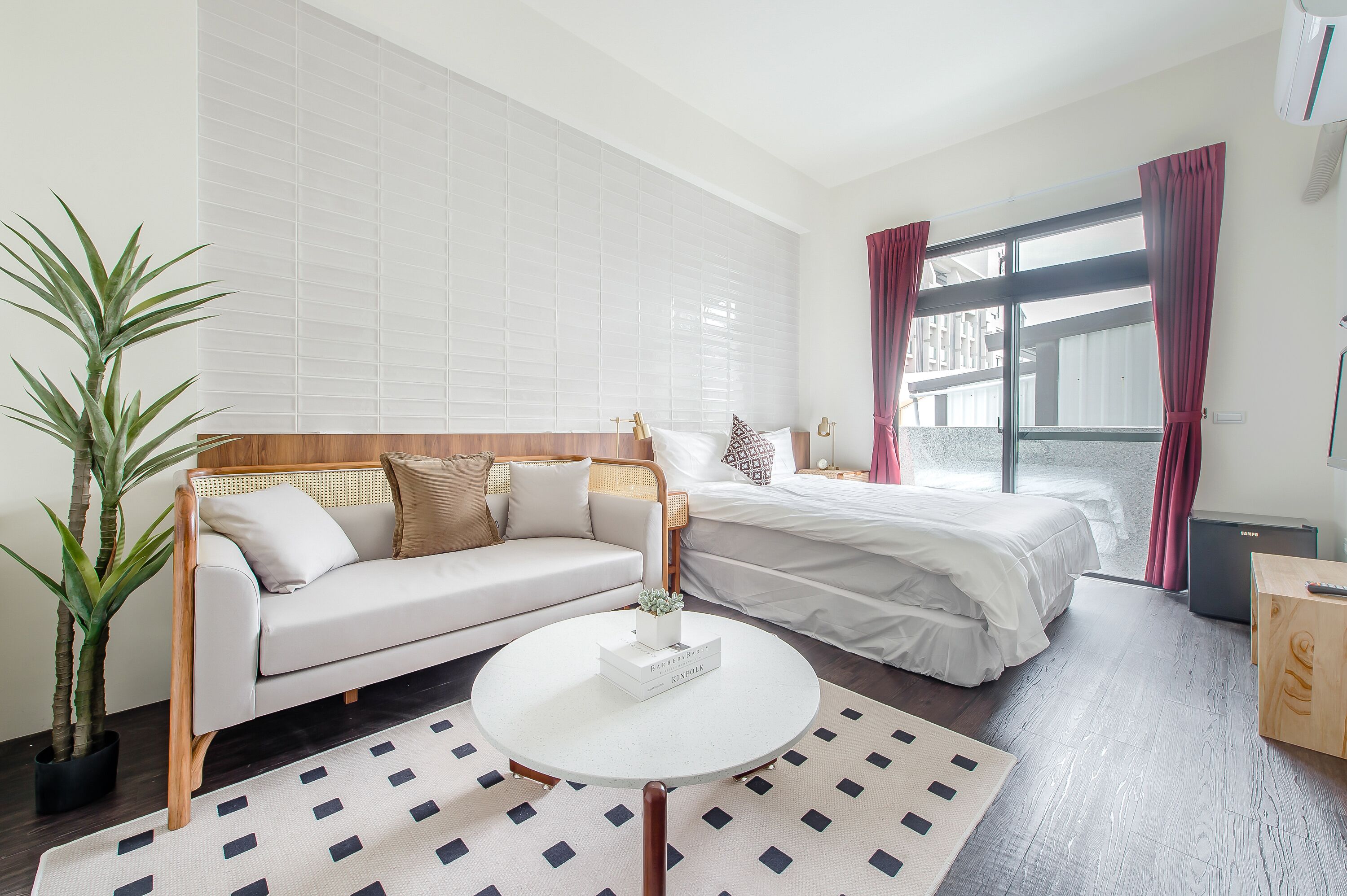 A modern hotel room with a white sofa, a bed with white bedding, a round coffee table, and a large window with curtains.