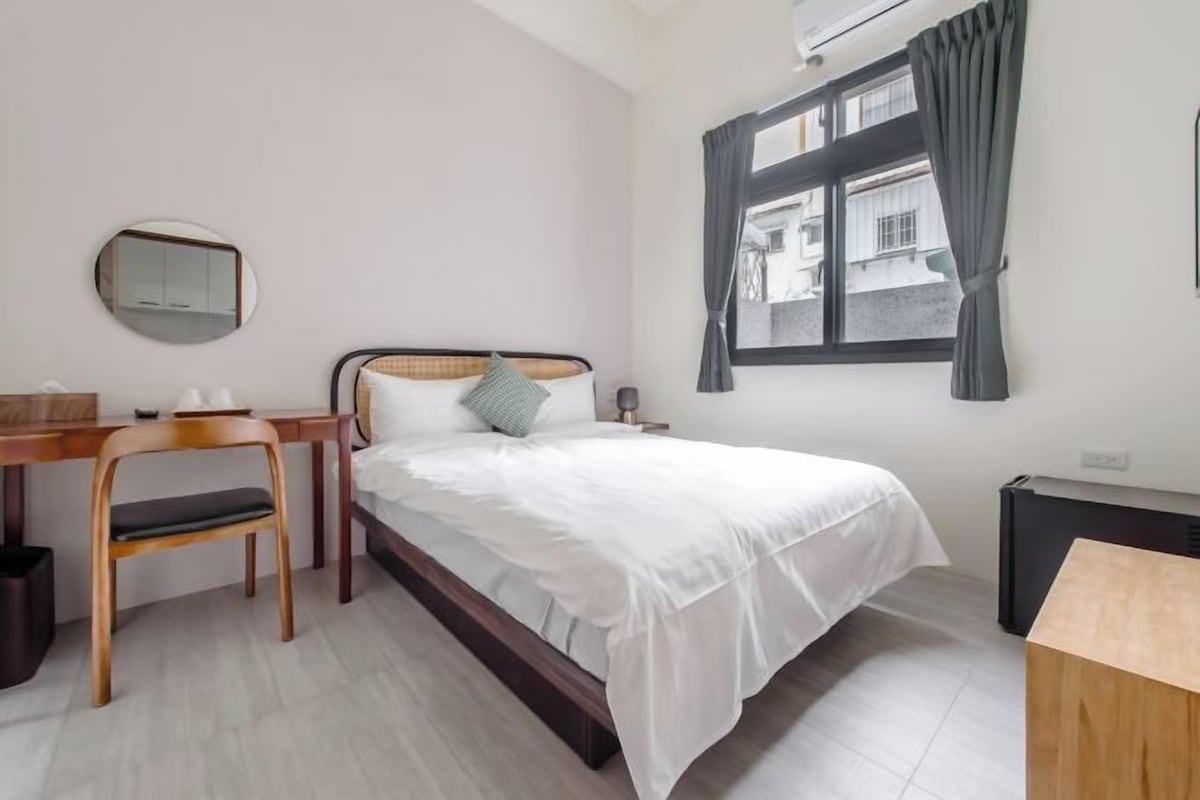 A bedroom with a bed, a desk, a chair, a mirror, and a window with curtains.