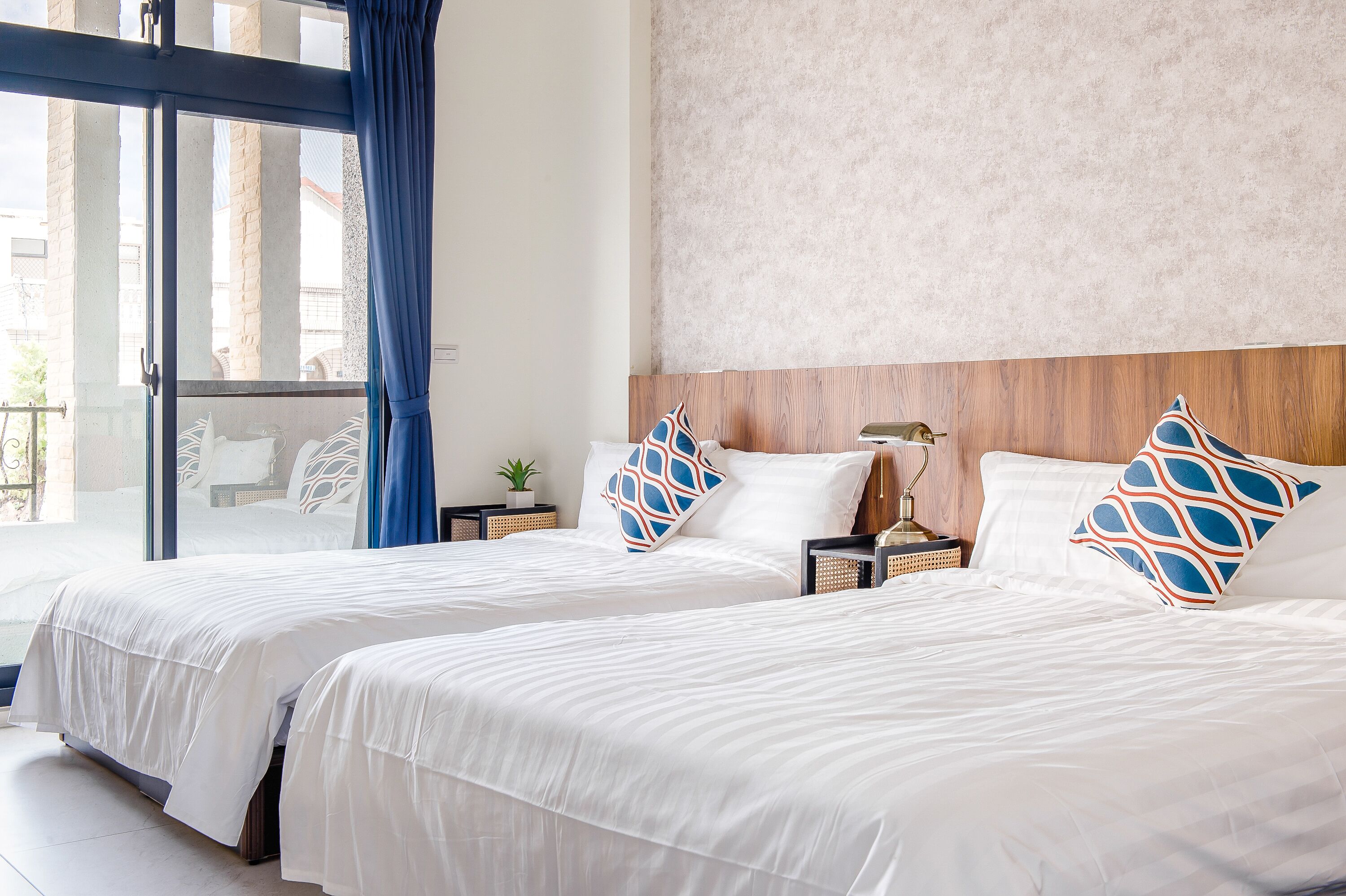 Habitación de hotel con dos camas, un ventanal con cortinas azules, un cabecero de madera y una mesita de noche con una lámpara y una planta.