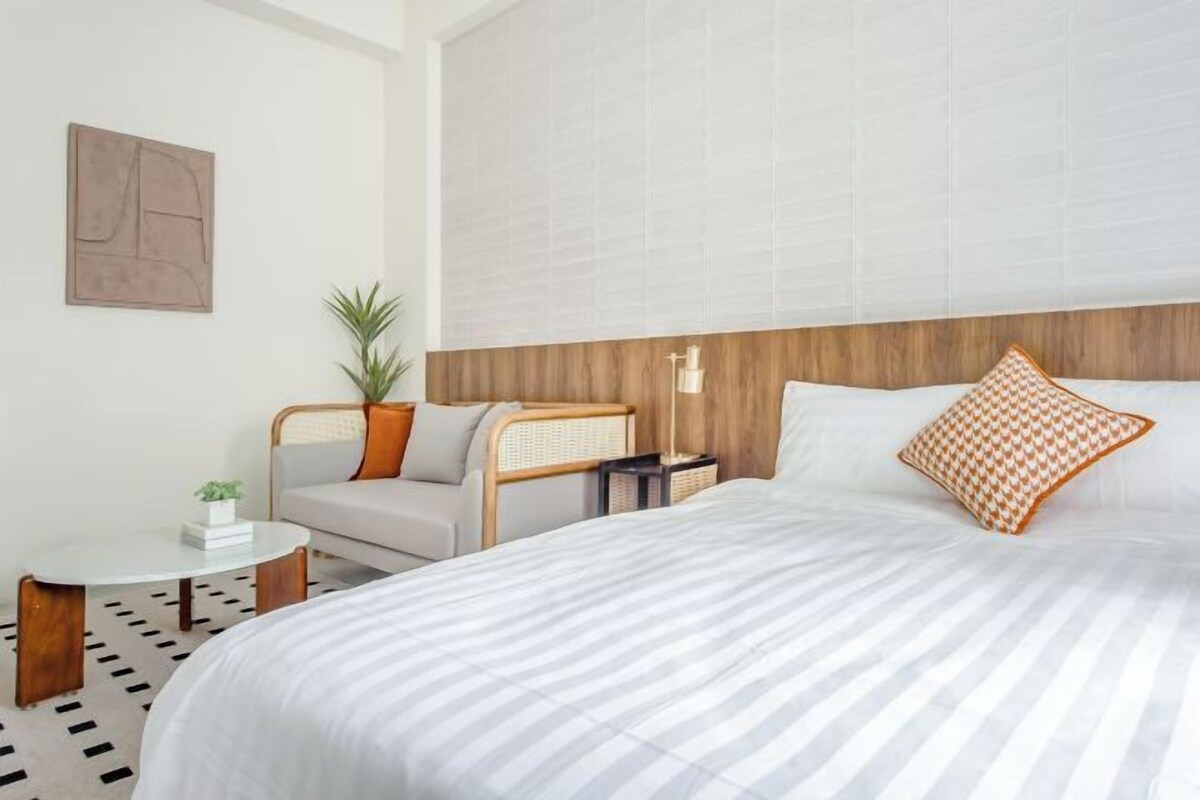 A hotel room with a large bed, a small round table, a chair, a potted plant, and a wall-mounted light fixture.