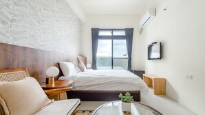 Ein modernes Hotelzimmer mit einem großen Bett, einem hölzernen Kopfteil, einem runden Couchtisch mit Pflanze und Blick zum Himmel durch das Fenster.