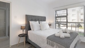 A modern bedroom with a large bed, two bedside tables, a grey headboard, and a view of the street through large windows. - Ballito Village 69 by HostAgents (Ballito)