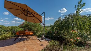 An outdoor patio with a table and chairs, a large umbrella, and lush greenery. - GuestHost - Cozy Apt with Terrace and Private parking! x4 (Gonars)