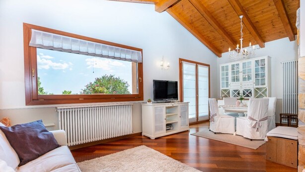 A living room with a large window, wooden ceiling, white furniture, and a TV. - GuestHost - Cozy Apt with Terrace and Private parking! x4 (Gonars)