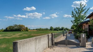 A gated entrance with a concrete barrier, a gravel path, and a building with a sloped roof. - GuestHost - Cozy Apt with Terrace and Private parking! x4 (Gonars)