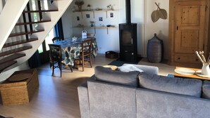 Una sala acogedora con un sofá gris, una mesa de comedor con sillas, una escalera, un estufa de leña negra y un armario de madera.