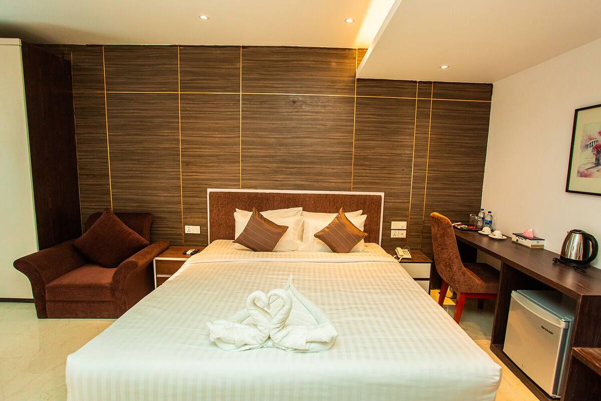 A modern hotel room with a large bed, two brown armchairs, a desk with a refrigerator, and a wall-mounted TV.