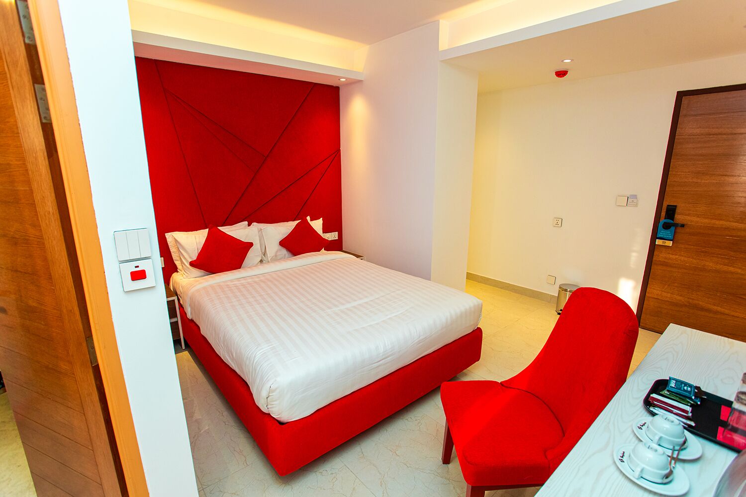 A modern hotel room with a red accent wall, a bed with white bedding, a red chair, and a small table with a tray of cups and a book.
