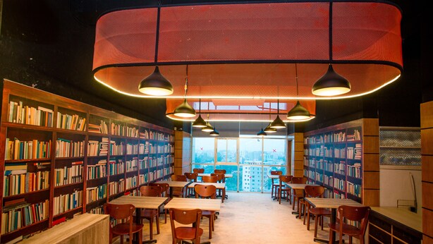 A library with bookshelves, tables, chairs, and large windows offering a city view. - Royal Raj Hotel & Condominium (Rajshahi)