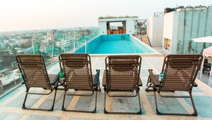 A rooftop pool area with three wicker chairs facing the pool, a view of the cityscape, and a staircase leading to an upper level. - Royal Raj Hotel & Condominium (Rajshahi)