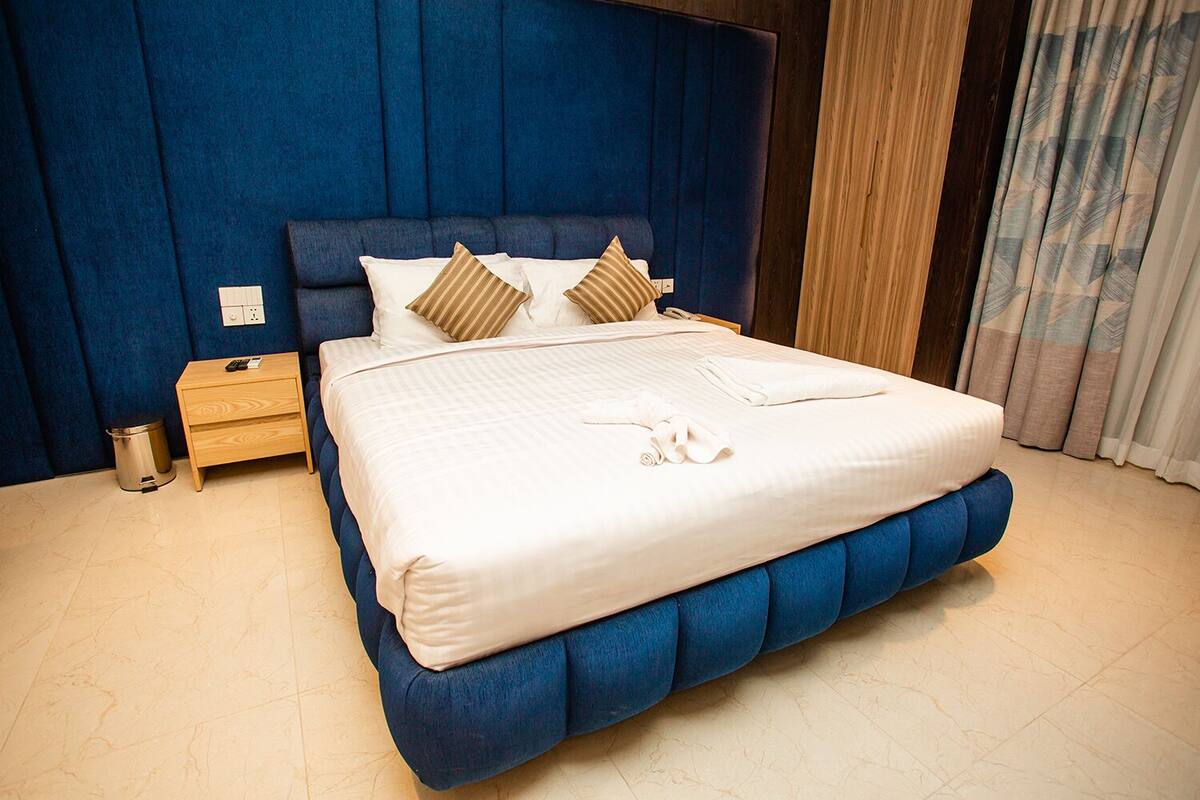A neatly made bed with a blue upholstered headboard, a wooden nightstand, and a small trash bin.