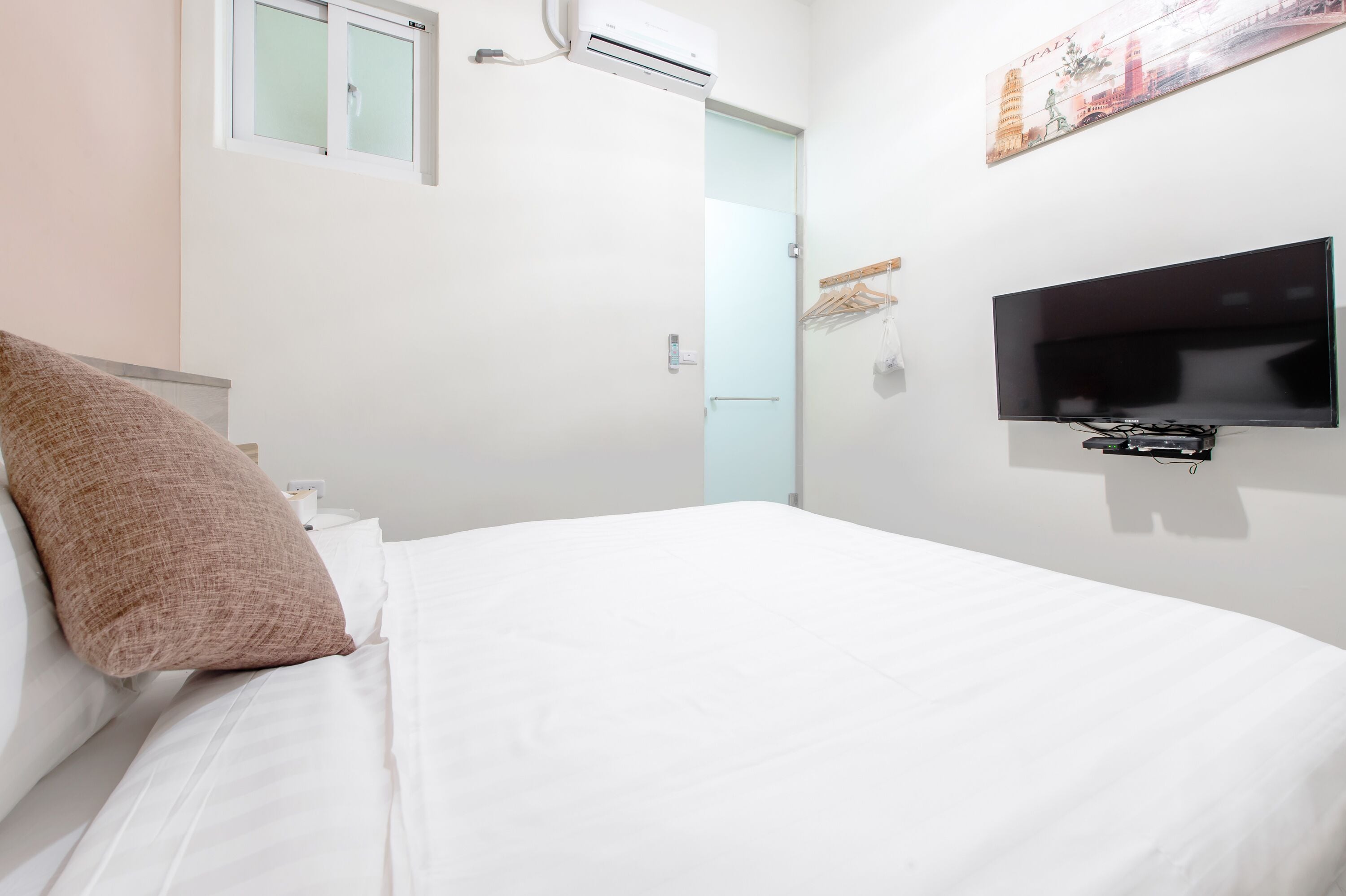 Ett hotellrum med en säng, en tv som är monterad på väggen, en luftkonditioneringsanläggning och ett fönster.