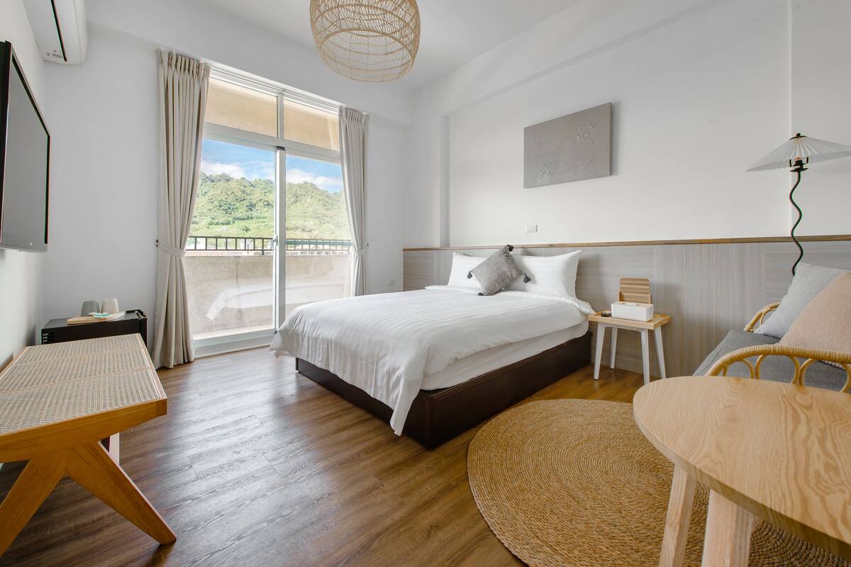 A bedroom with a bed, a small table, a chair, a bedside table, and a view of greenery outside.