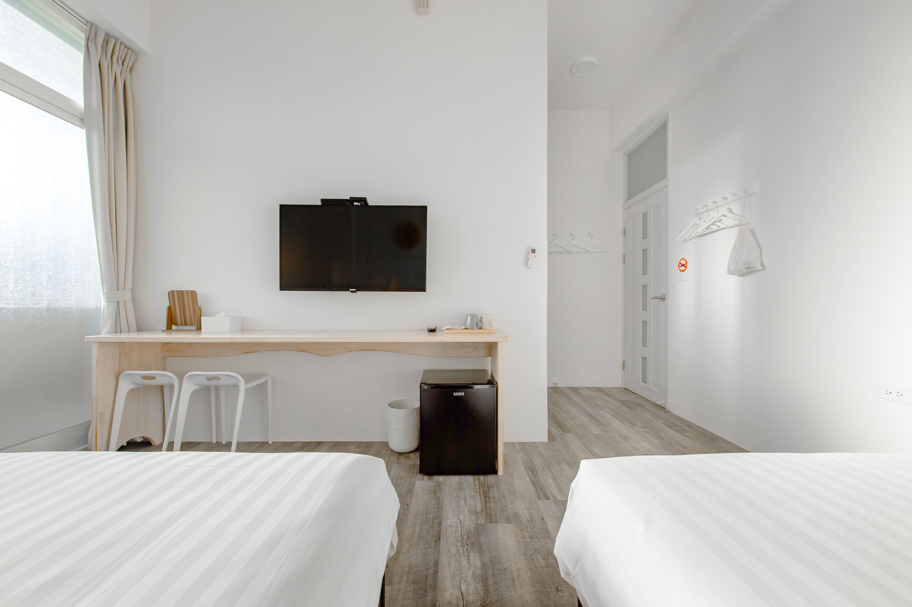 A hotel room with a wooden desk, two white chairs, a TV mounted on the wall, and two beds.