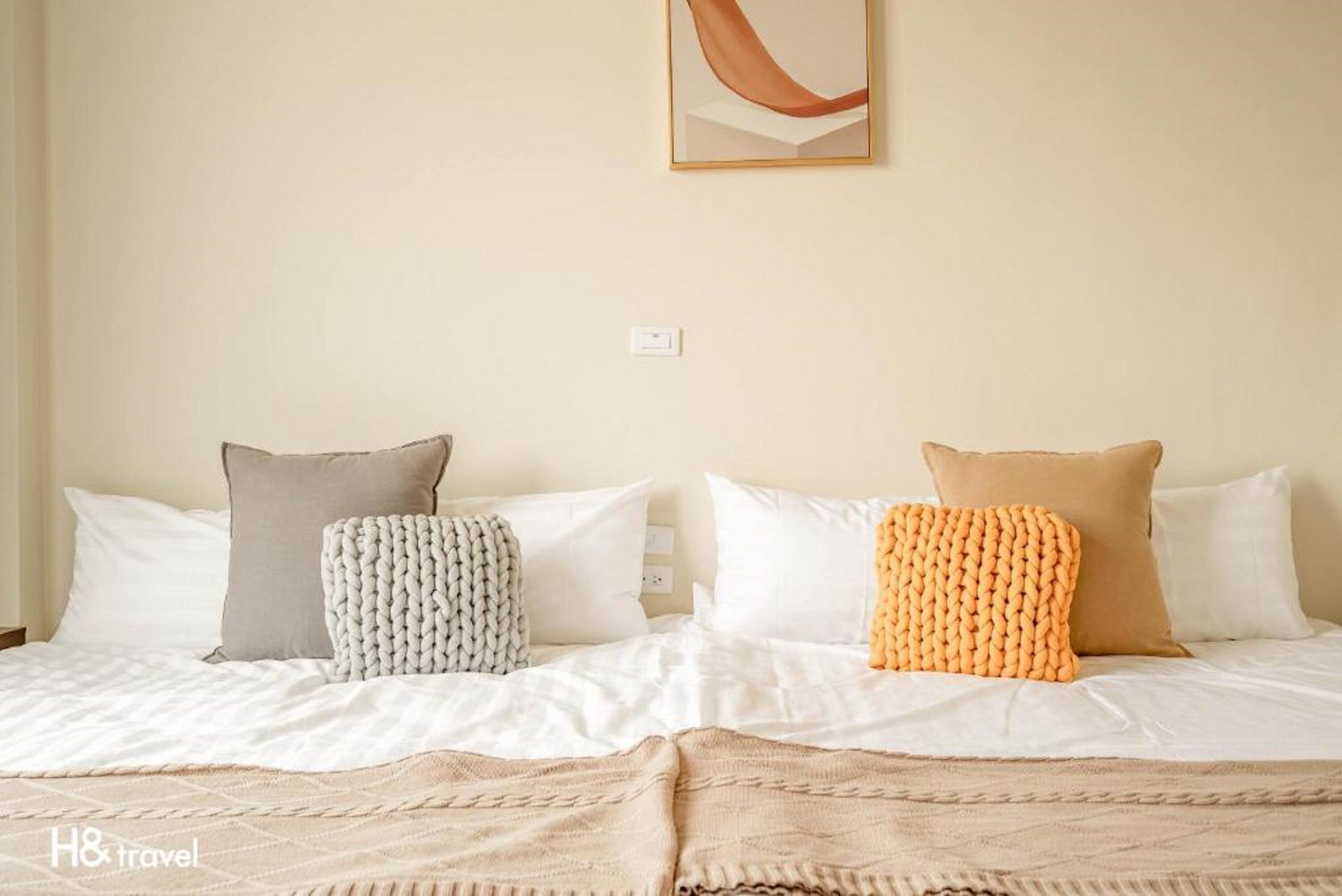 A double bed with white bedding and two decorative pillows, one grey and one orange, in a hotel room.