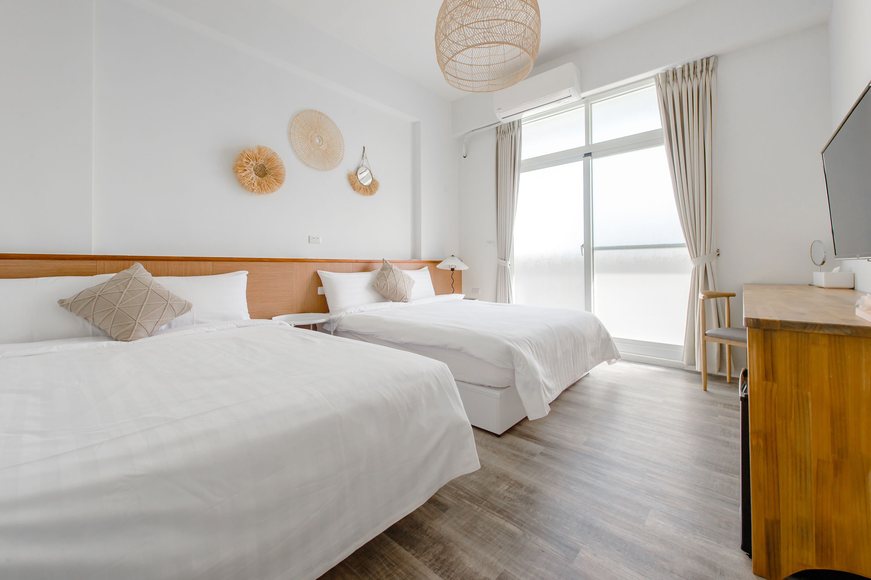 A hotel room with two beds, a wooden headboard, a desk, a chair, a TV, and a large window with curtains.