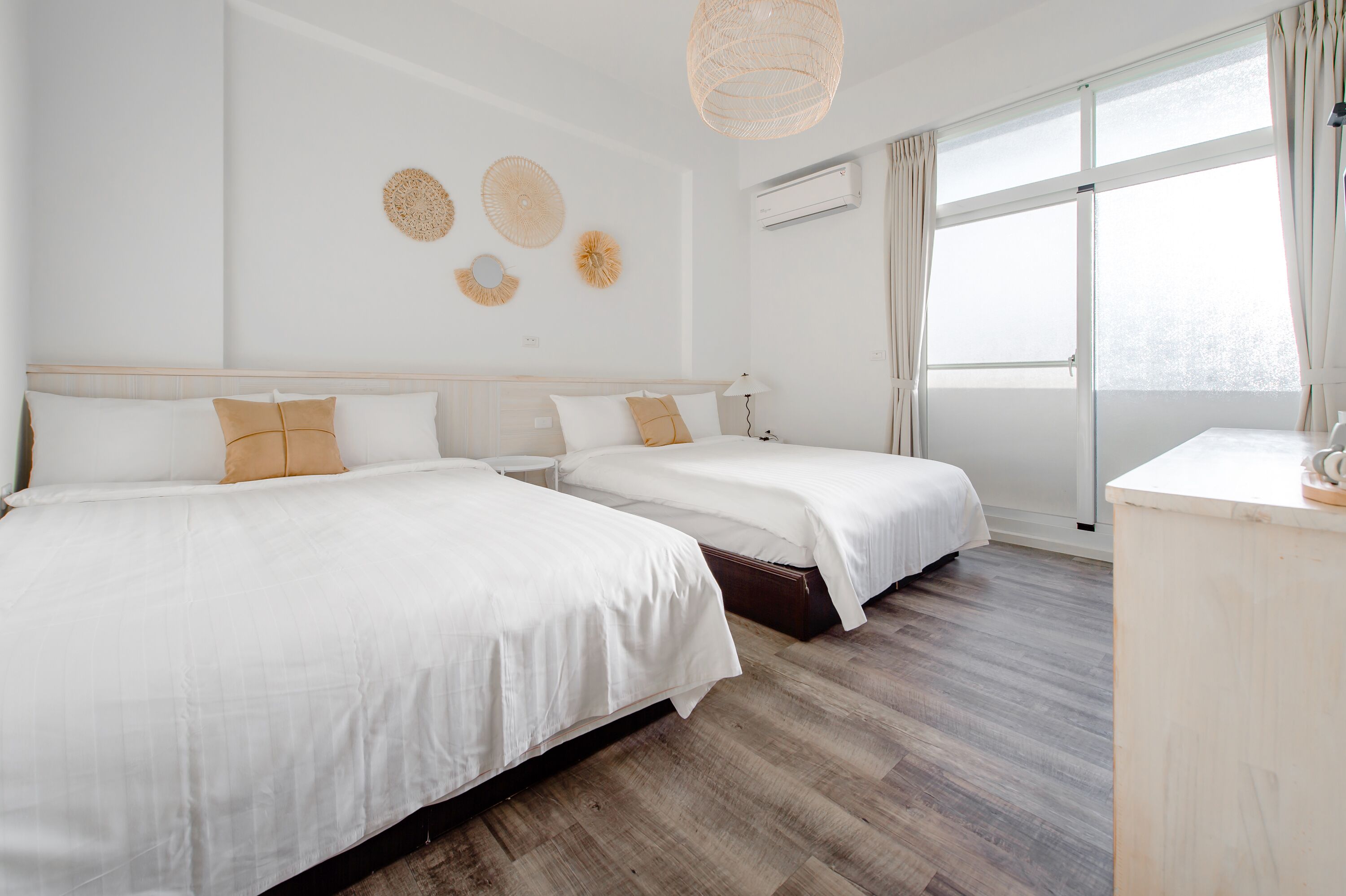 A hotel room with two beds, a wooden floor, a large window with curtains, and wall decor.