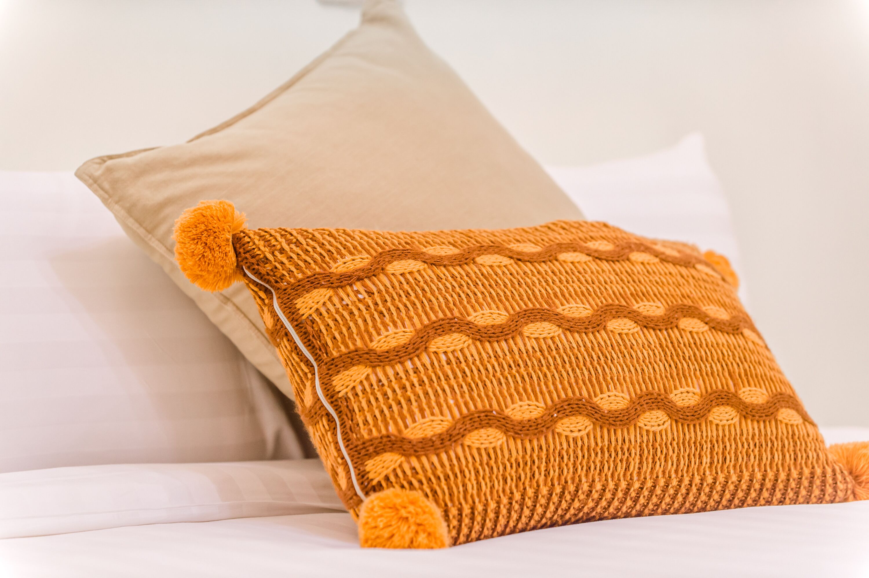 Una cama con ropa de cama blanca y una almohada anaranjada de punto con pompones.