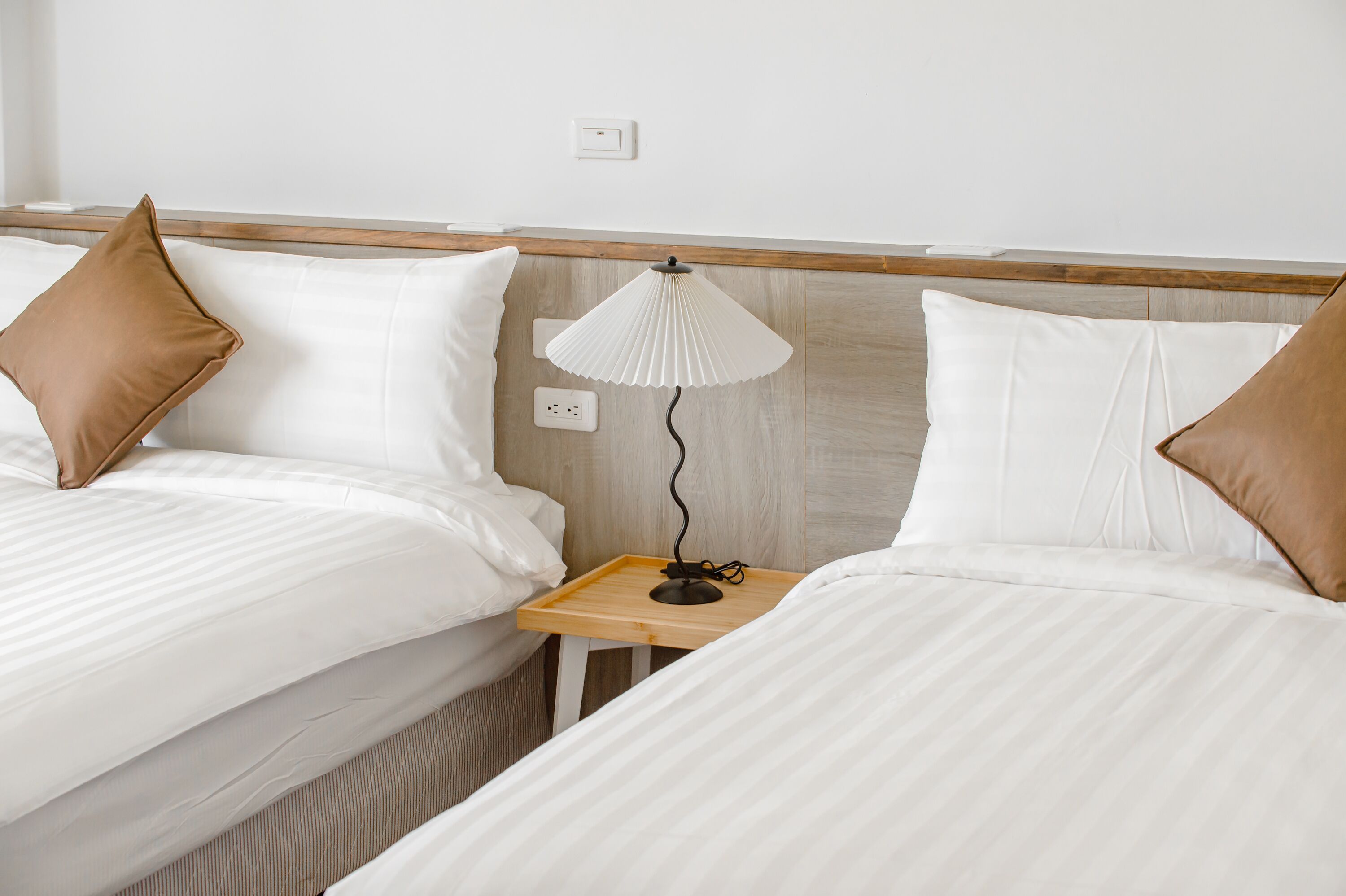 A hotel room with two beds, each with white bedding and brown pillows, a wooden nightstand with a lamp, and a light switch on the wall.