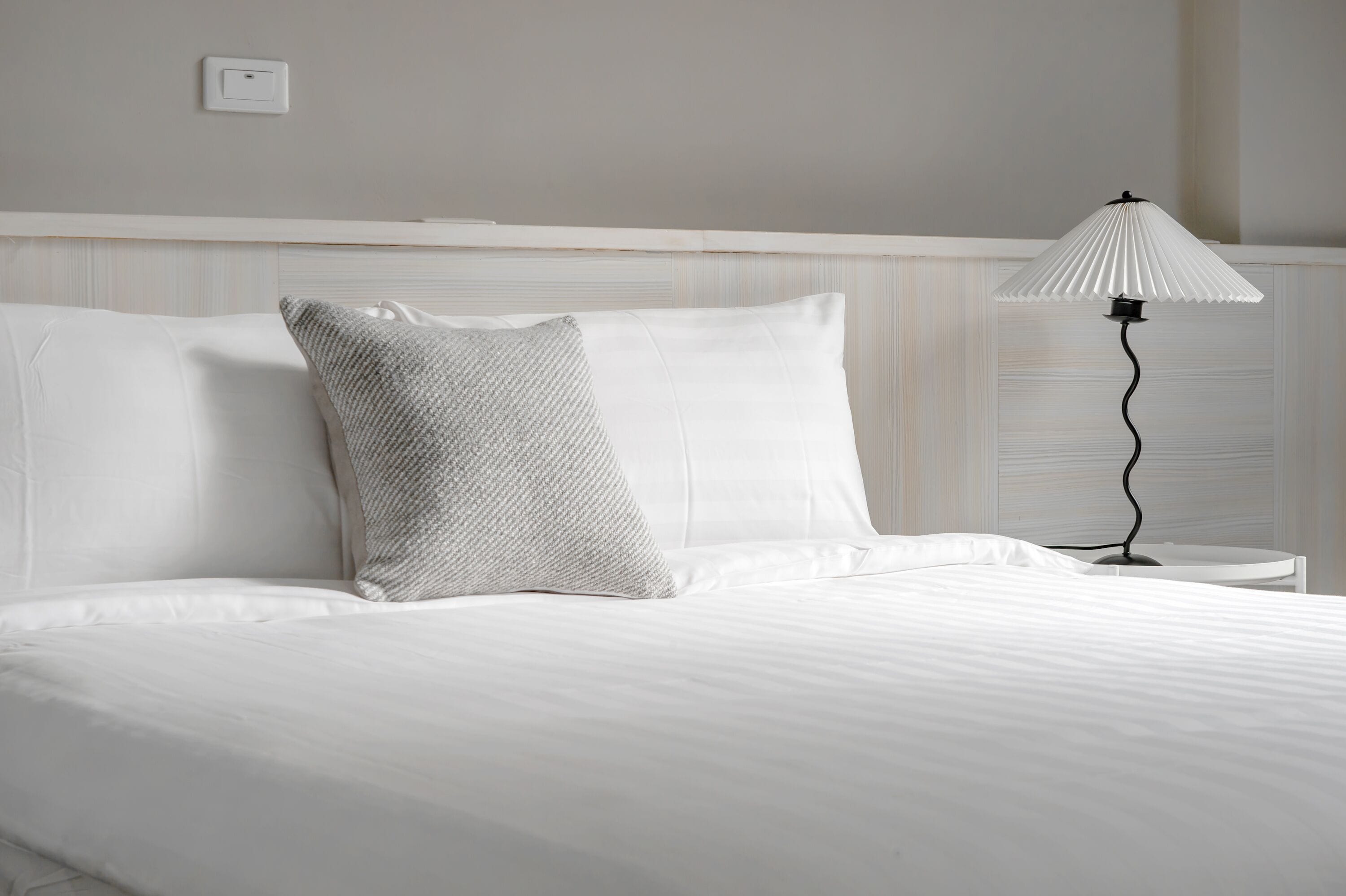 Una cama bien hecha con sábanas blancas y un cojín con textura, una lámpara de noche y un interruptor de luz en la pared.