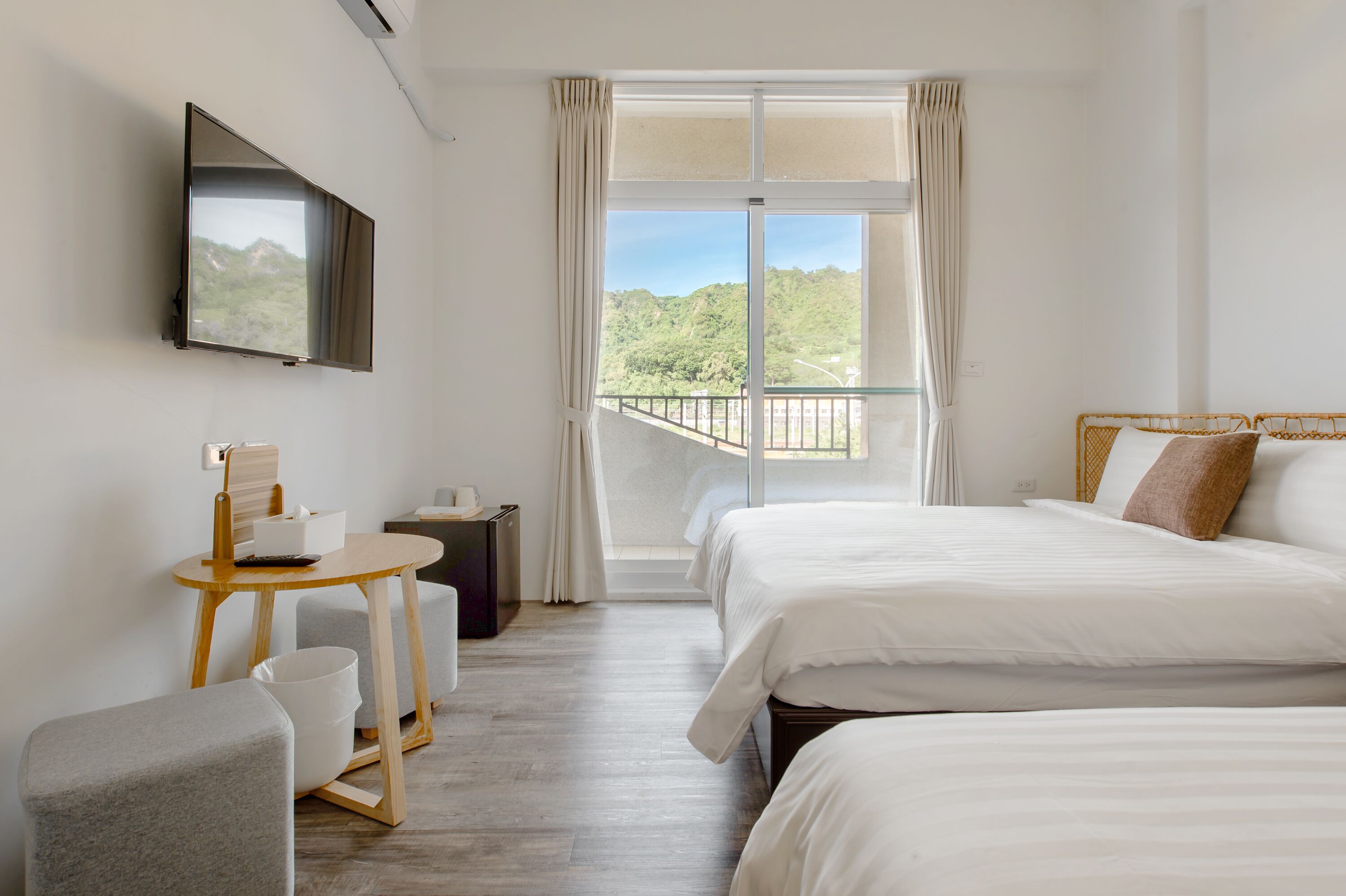 A hotel room with two beds, a TV mounted on the wall, a small table with a chair, and a window with a view of greenery.