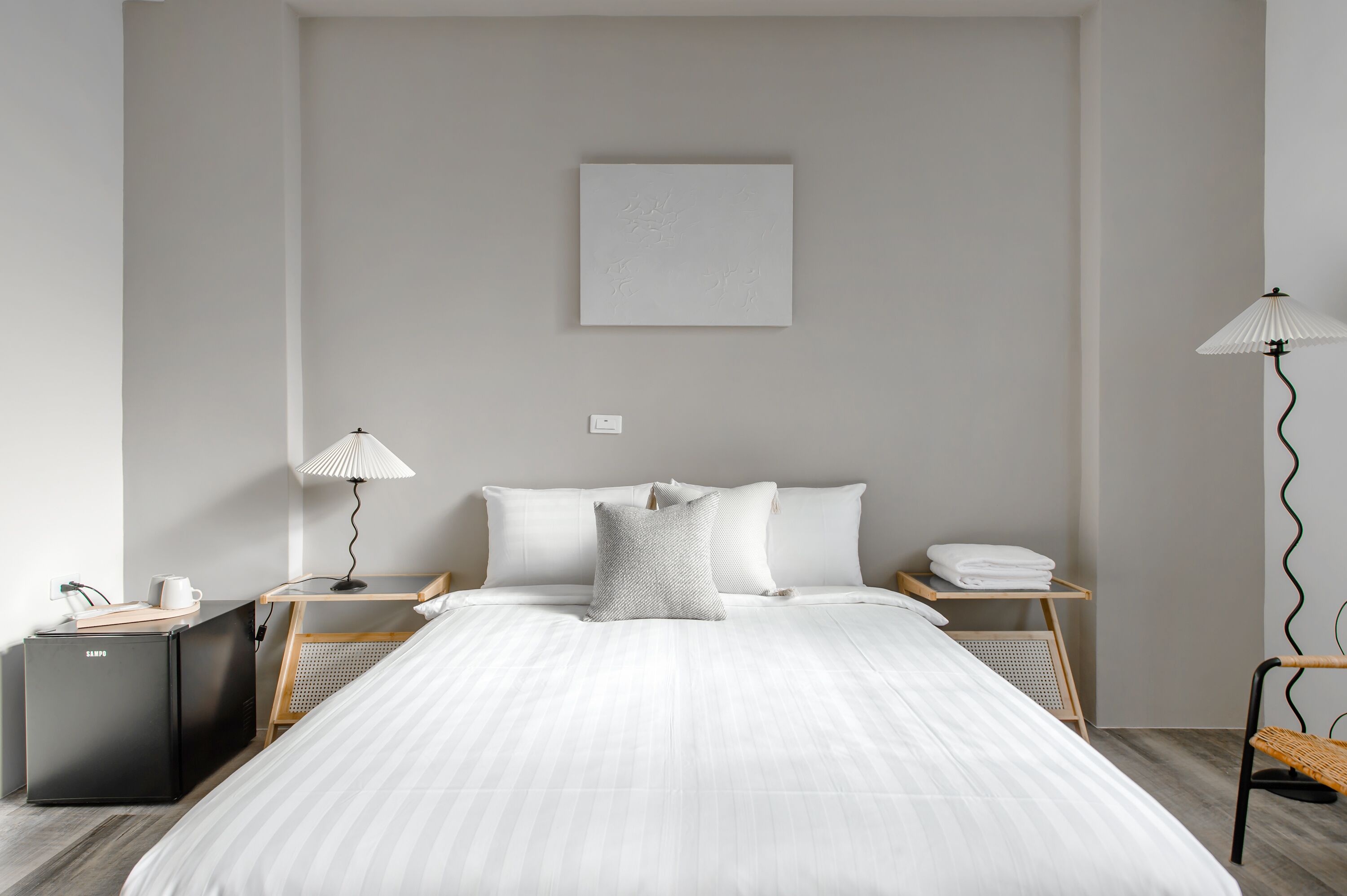 A modern hotel room with a large bed, a small desk with a lamp, a bedside table with a lamp, and a small refrigerator.
