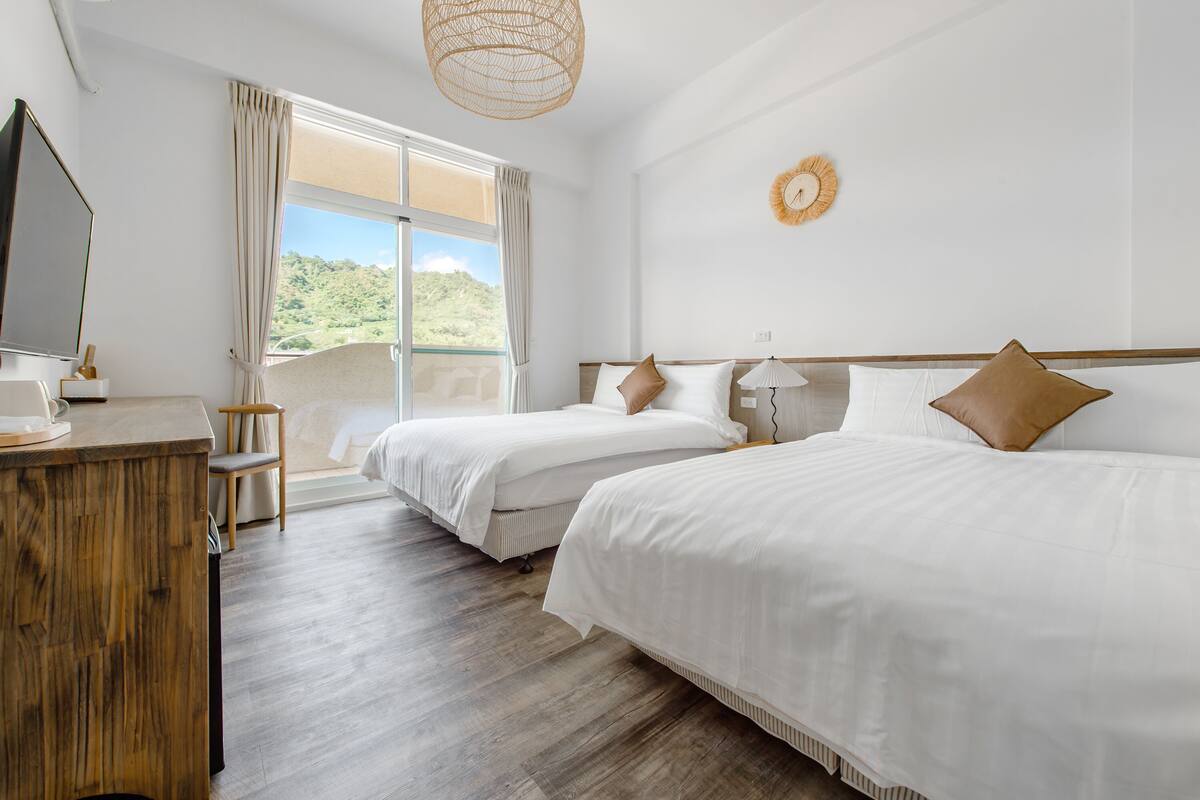 A hotel room with two beds, a wooden desk, a television, and a view of greenery through the window.