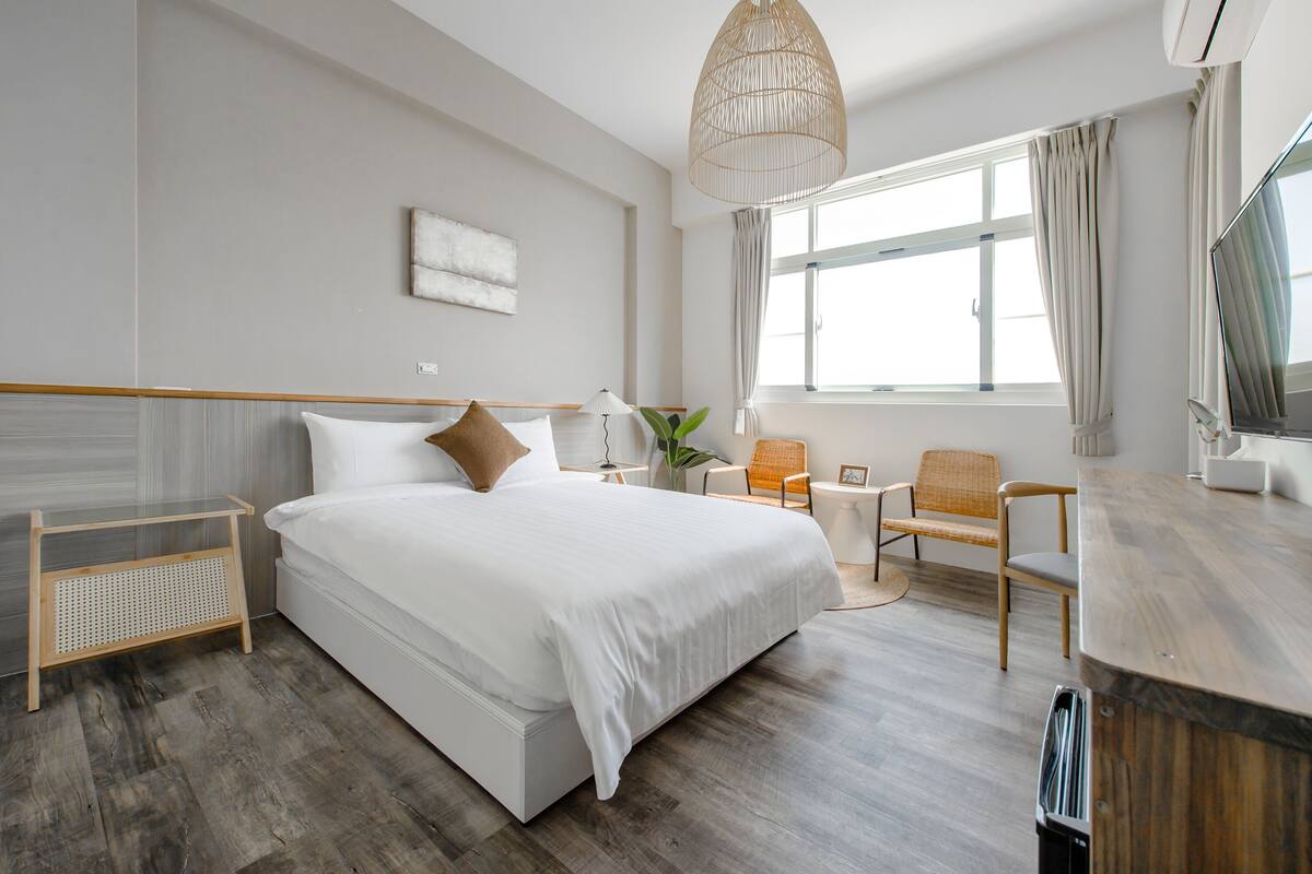 A modern hotel room with a large bed, a wooden side table, a chair, a desk, and a large window with curtains.