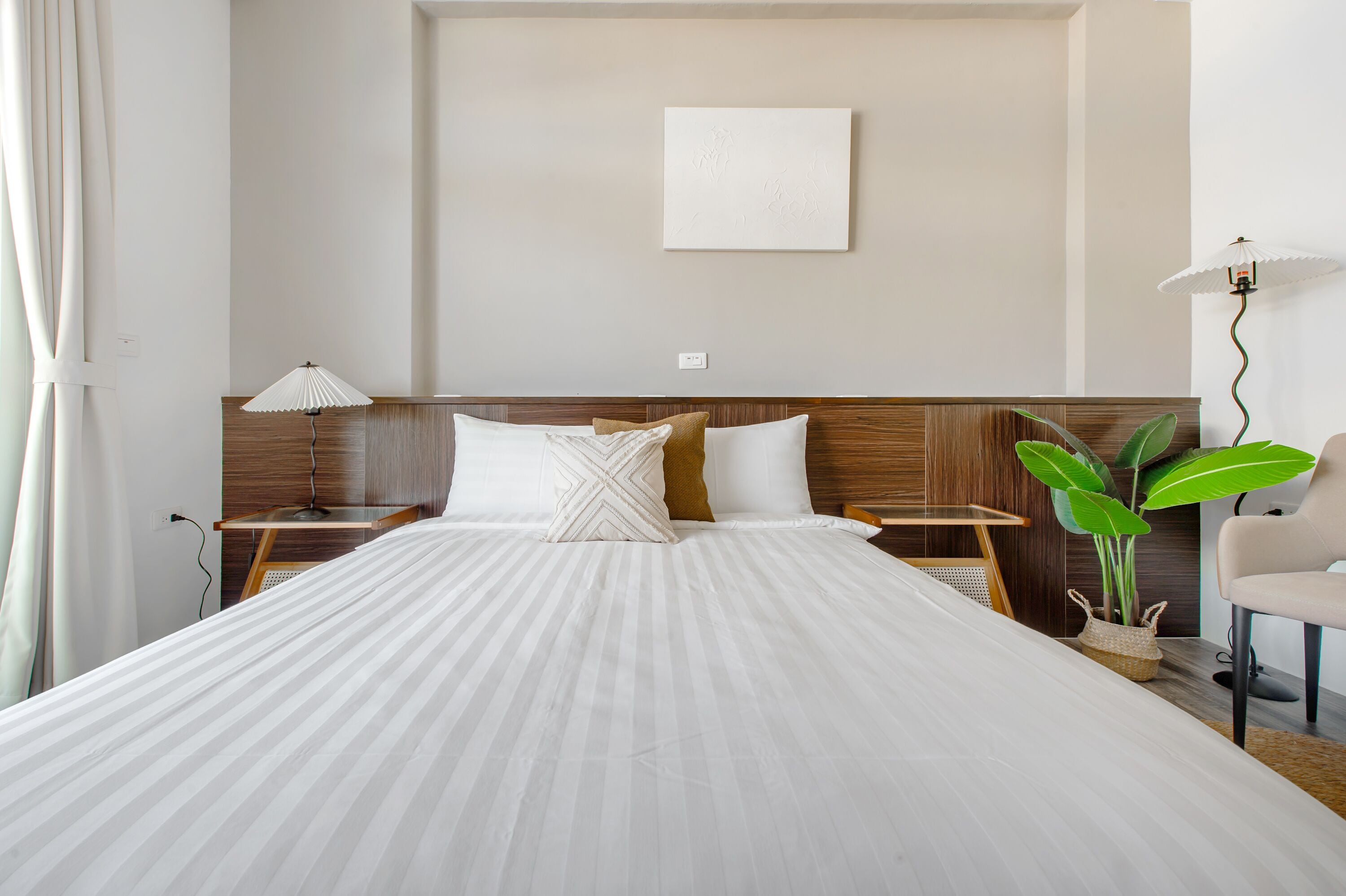 A neatly made bed with a wooden headboard, a bedside table with a lamp, a chair, and a potted plant.