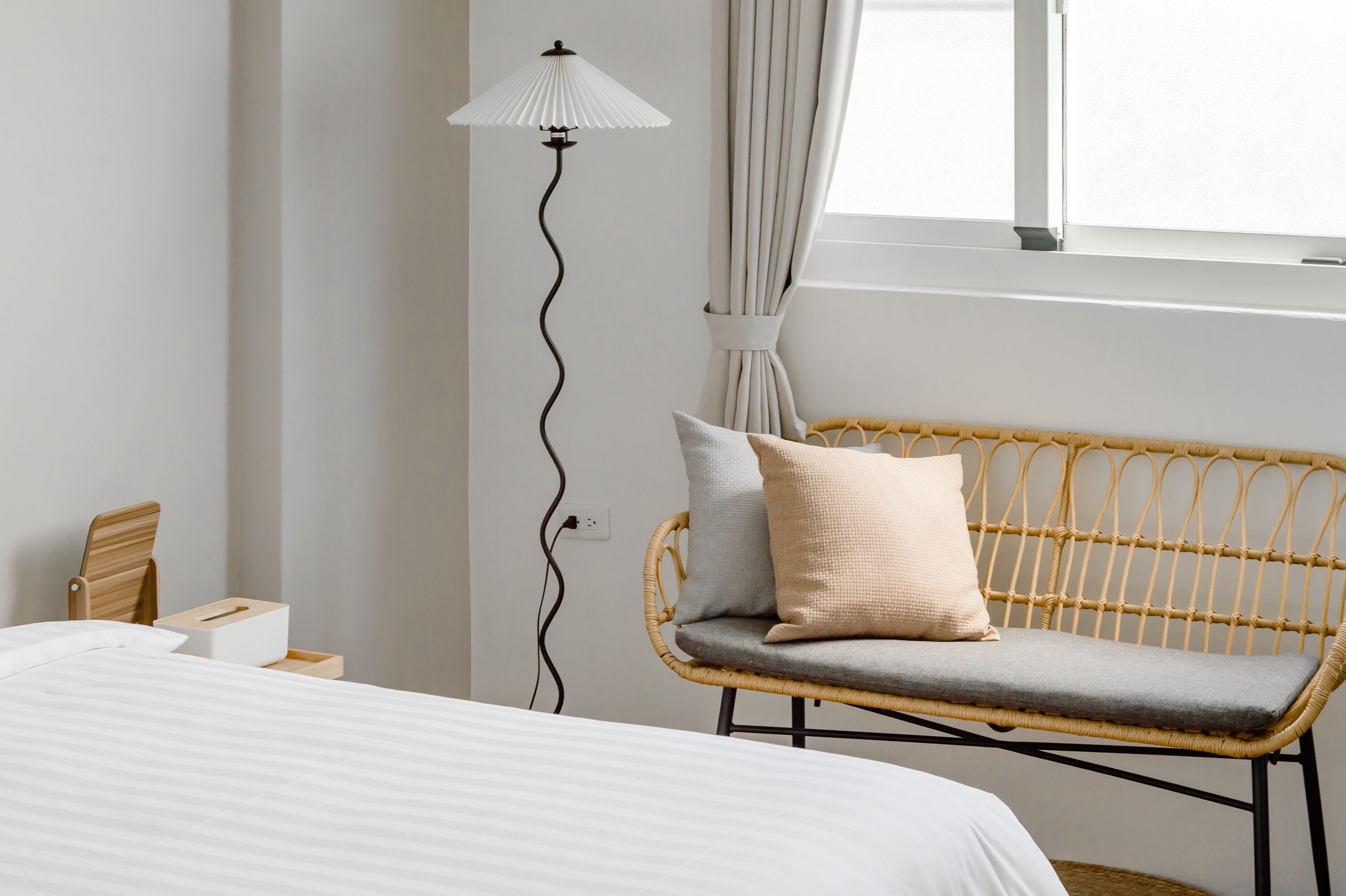 A bedroom with a bed, a wicker chair with cushions, a floor lamp, and a window with curtains.