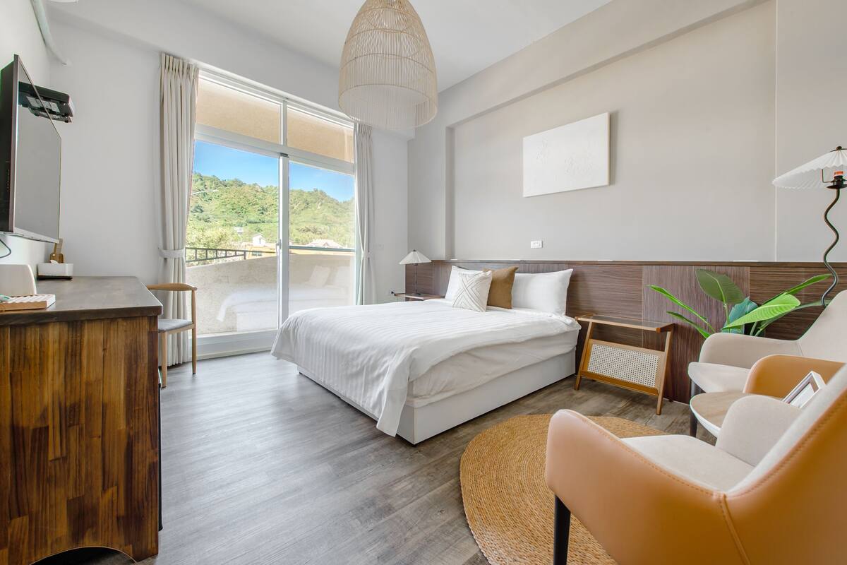 A modern hotel room with a large bed, a wooden desk, a comfortable armchair, and a view of the outdoors.