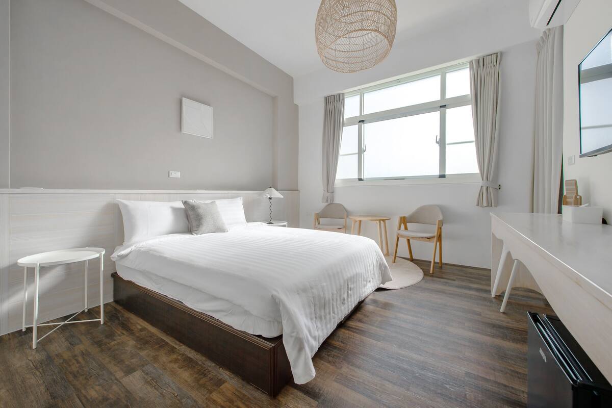 A bedroom with a bed, bedside tables, a desk, a chair, and a large window with curtains.